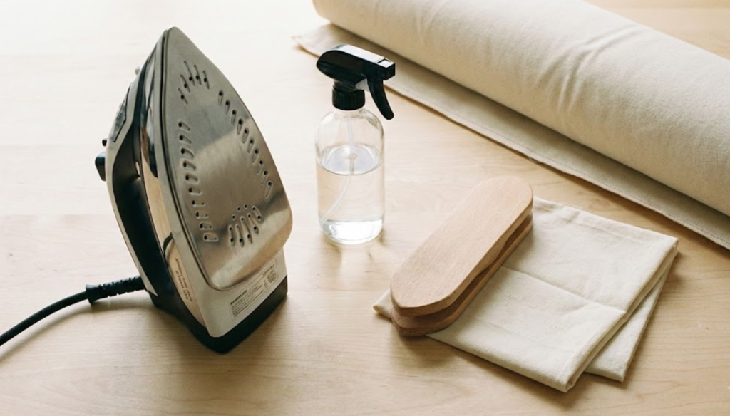 Essential tools for ironing heavy duck fabric including a steam iron, water spray bottle, wooden tailor's clapper, and muslin pressing cloth.